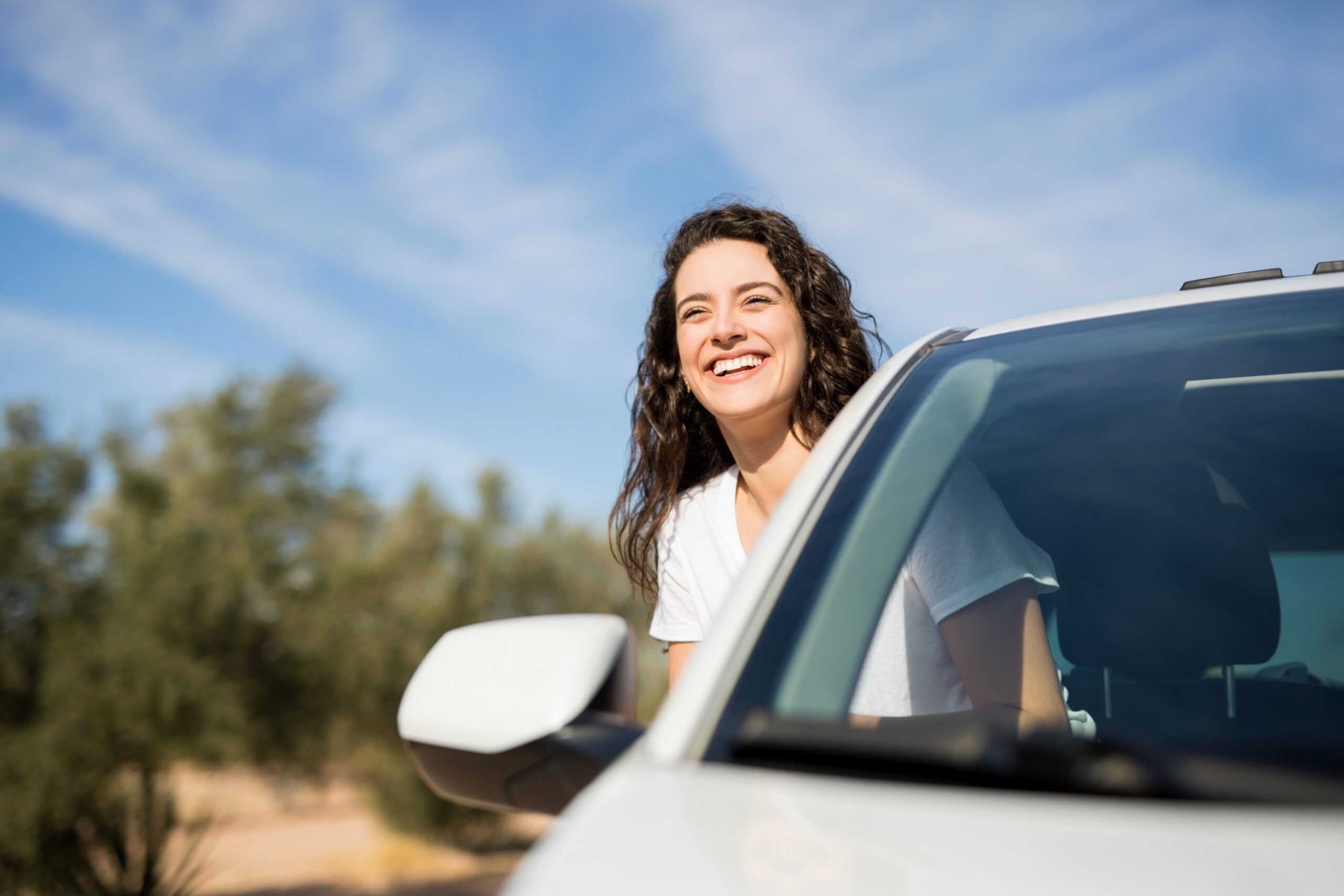 Portrait of smiling young woman sitting in car passenger seat with her head out of window looking the road and smiling